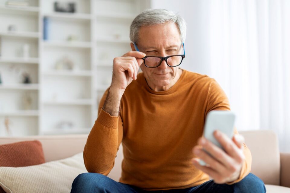 An older man squinting at a smartphone while lifting glasses in East Stroudsburg, PA